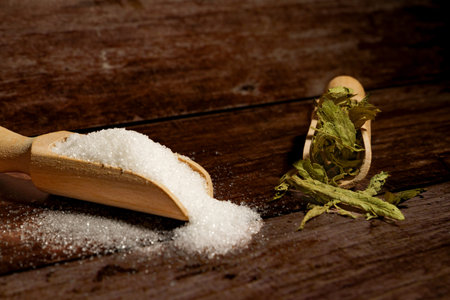Stevia leaves and white sugar granules in wooden scoops, offering natural and artificial sweetener choices on a dark wood backgroundの写真素材
