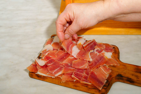 Hand picking thin, savory slices of cured serrano ham from a rustic wooden cutting board, ready to serve as a gourmet tapas or appetizer in traditional Spanish styleの写真素材