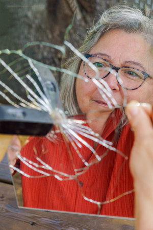 Mature woman with gray hair and glasses looking into a shattered handheld mirror, contemplating her...