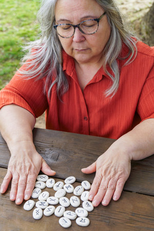 Elder woman with gray hair and glasses sitting at a wooden table outdoors, concentrating on white...