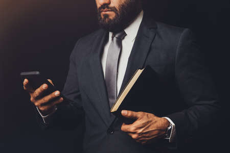 man in suit holding a bible in his arm and with the cell phone in handの写真素材