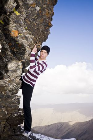 Young model climbing a small cliff at high altitude in casual clothes.