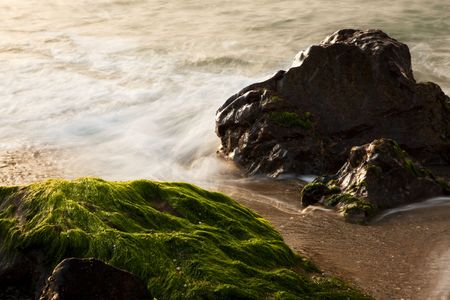 Long exposure on shore, blurred waves over green rocks.