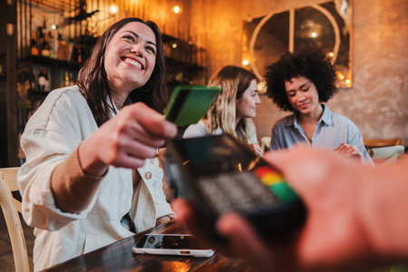 Happy young woman paying bill with a contactless credit card in a restaurant. Female smiling holding a creditcard and giving a payment transaction to the cashier.の写真素材