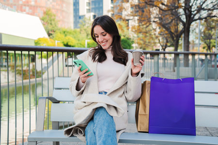 Happy young woman sitting on a bech using a smartphone to text messages after shopping on-line. Brunette female looking to the cellfone device sharing content on social media.の写真素材