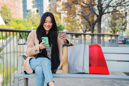 Young carefree asian woman typing message using a cellphone and taking a coffee after shopping. Teenager texting with smartphone app sitting on a bench outdoors. Communication conceptの写真素材