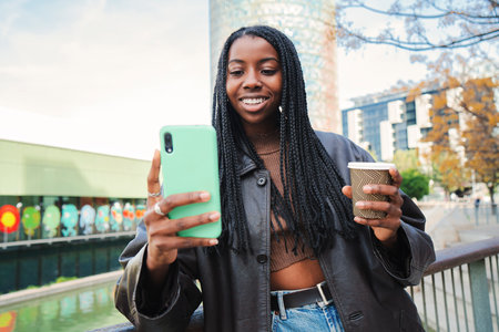 Young african american woman smiling and typing with cellphone app. Happy teenage girl sending messages using an smart phone after shopping outdoors. Communication concept. Slow motionの写真素材
