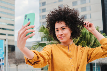 Young latin woman doing a selfie portrait with a cellphone touching her curly hair. Happy hismanic...