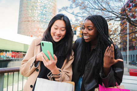 Two multiracial young women smiling watching a funny video on a cellphone. A couple of multiethnic girls with surprised face looking for a sale on internet using a smartphone outdoors.の写真素材