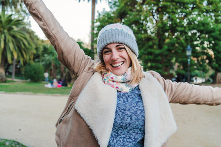 Front view of one happy caucasian blonde young woman smiling with white perfect teeth, wearing a beanie hat and looking excited to the camera in a park outdoorsの写真素材