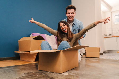 Young caucasian happy couple having fun playing with cardboard box in their new house after moving. Husband and wife arriving at their new home with packages enjoying the change . Loan conceptの写真素材
