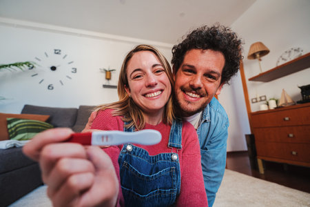Happy caucasian young couple smiling excited showing at camera their positive pregnancy test at home. Husband and wife celebrating that they are expecting a baby holding a predictor in their hands.の写真素材