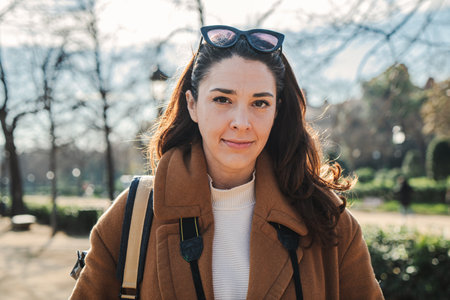 Close up portrait of young caucasian woman looking pensive at camera. Front view of adult business brunette female standing at park outdoors. City life concept.の写真素材