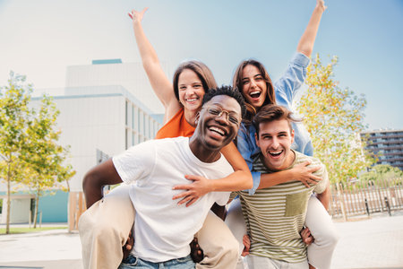 Two multiracial young friends giving piggybacks ride to his girlfriends. A group of happy people having fun and smiling at weekend activity. Men carrying women on his shoulders looking at cameraの写真素材