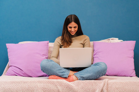 Young caucasian woman working with laptop computer sitting on a couch in front of blue wall. Happy beautiful female smiling using a notebook with wireless internet connection at home. Domestic lifeの写真素材