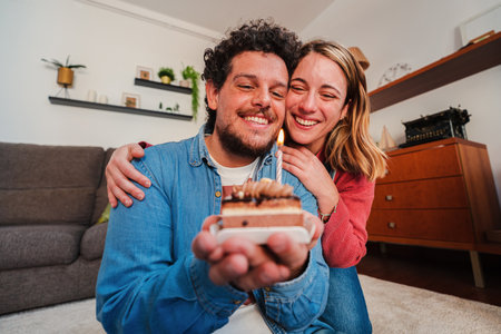 Happy Young couple celebrating a birthday with a little cake blowing the candle. Blonde wife giving a funny surprise to her husband for their anniversary while hugging him from behind at home.の写真素材