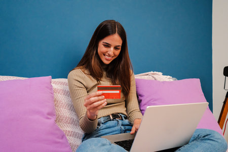 Young caucasian brunette woman booking a room using a laptop computer and paying with credit card sitting on a couch at home indoors. One female purchasing online on internet. Finance concept.の写真素材