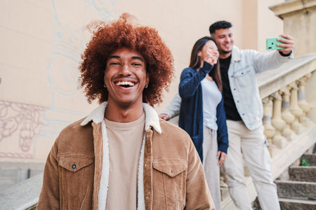 On foreground a handsome happy african american young man smiling and looking at camera with afro hair, at background two friends taking a selfie portrait. front view of teenage toothy male laughingの写真素材