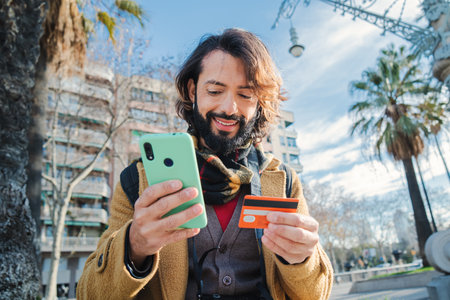 young man paying a purchase with a credit card and holding a smart phone device standing outdoors. Happy bearded male using a mobilephone app for purchase a delivery and look the finance banking.の写真素材