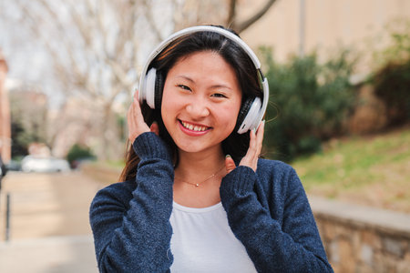 One chinese young woman listening music with headphones using wireless connection. Happy asian girl wearing earphones tu hearing songs using streaming platform app with cellphoneの写真素材