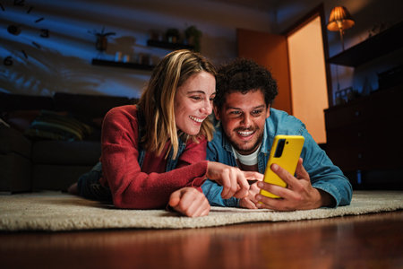 Couple watching funny videos together on a cellphone app lying on the floor at home. Young caucasian wife and husband on the carpet browsing on internet using a smart phone at late night.の写真素材