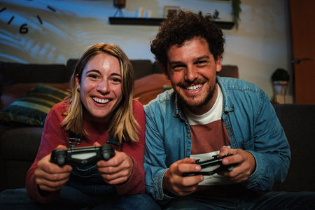 Young couple having fun plaing video games with a console together sitting at couch at home. Two happy friends smiling and gaming at night holding the controller and touching gamepad buttonsの写真素材