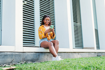 African american female student smiling using a cellphone, reading awesome news or shopping online enjoying her free time sitting at university campus . Happy teenage womanの写真素材