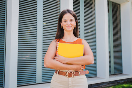 Portrait of a beautiful young caucasian female student holding notebook in hands looking at camera at university campus outdoors. Front view of a hispanic happy teenage woman withの写真素材