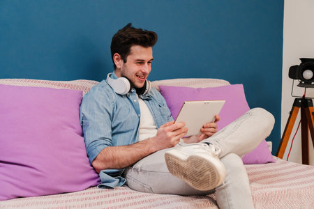 Young man using a tablet app to browse on internet sitting at home couch. Male teenager smiling and touching the screen of his device to do a delivery order resting at herの写真素材