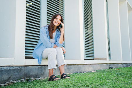 young caucasian woman smiling and talking by mobile phone. Lady having a friendly call conversation by smartphone resting outside. Joyful teenage female student using a cellphoneの写真素材