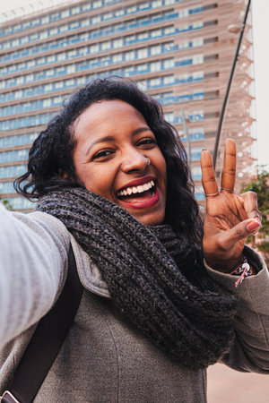 Vertical portrait of carefree woman looking at camera and smiling while taking a selfie and showing peace sign outdoors. Young african american female laughing with perfect whiteの写真素材