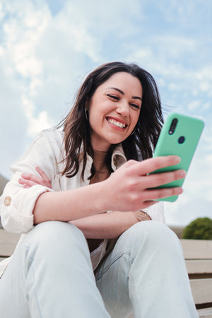 Vertical portrait of young adult woman watching the social media on a cellphone app sitting outdoors. Carefree female smiling and having fun texting messages with a smartphoneの写真素材