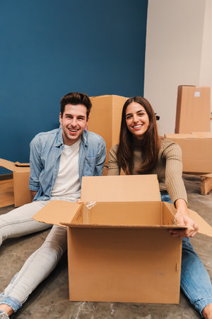 Vertical. Young caucasian couple smiling moving to their new home, using cardboard boxes to carry their things. Husband and wife sitting on the floor of their apartment unpackingの写真素材