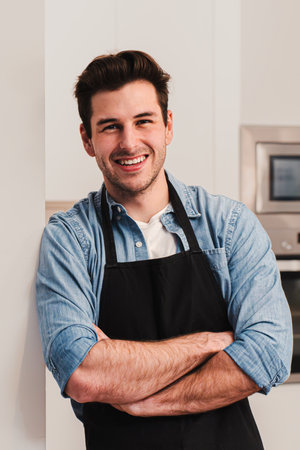 Vertical portrait of handsome caucasian man wearing an apron standing in the kitchen, smiling and...