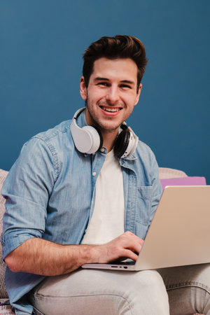 Vertical portrait of handsome entrepreneur freelancer man working at home with a laptop sitting on a couch smiling and looking at camera. Young guy browsing on internet using aの写真素材