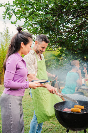 Vertical portrait. Middle adult happy couple cooking meat on a barbecue bbq party with their friends at home backyard. Husband and wife grilling food together on barbeque weekendの写真素材