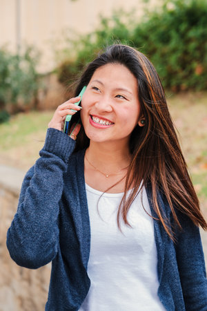 Vertical portrait of a teenage female student smiling and having a cellphone call conversation. Happy young chinese woman talking by smartphone outside. Gossip lady using a mobileの写真素材