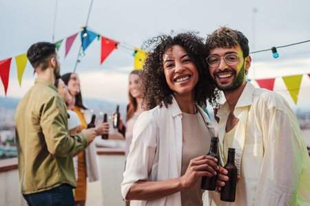 Portrait of young adult multiracial couple smiling, laughing and looking at camera holding beer bottles. Two people staring front on a party celebration with their best friendsの写真素材