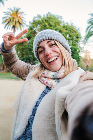 Vertical individual portrait of one happy caucasian blonde girl smiling taking a selfie portrait with a cellphone. European happy young woman doing the peace sign with the fingersの写真素材