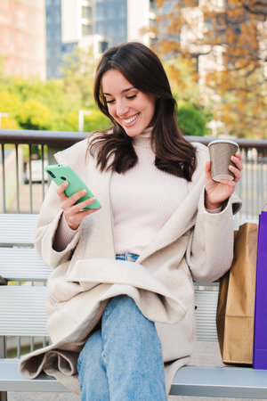 Vertical portrait of a happy young woman sitting on a bech using a smartphone to text messages after shopping on-line. Brunette female looking to the cellfone device sharingの写真素材