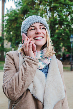 Vertical individual portrait of happy blonde girl having a mobile phone call at park outdoors. Young european woman smiling with perfect white teeth talking by cellphoneの写真素材