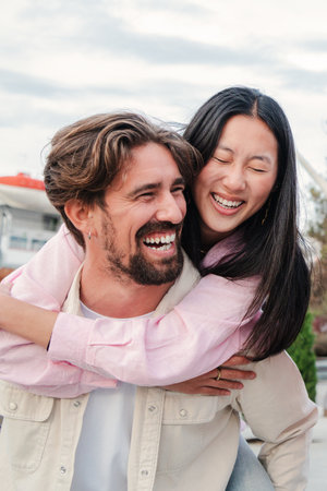 Vertical portrait of a young adult multiracial married couple having fun. Caucasian boyfriend giving a piggyback ride to his asian girlfriend on a weekend. Husband carrying onの写真素材