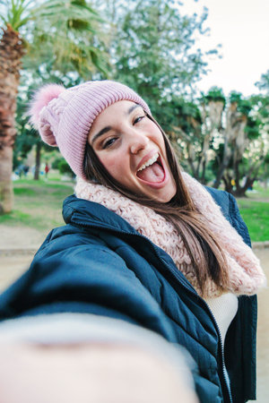 Vertical portrait of a young caucasian woman smiling taking a selfie in a park outdoors with scarf, beanie hat and autumnal clothes. Point of view of a teenage girl doing a photoの写真素材