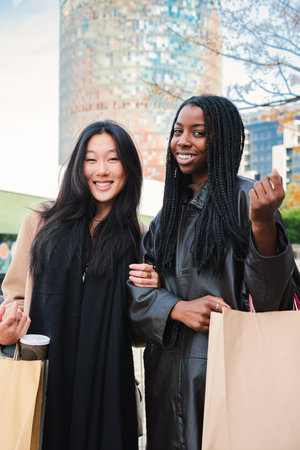 Vertical portrait of two happy young women smiling and looking at camera, carrying shopping bags. A couple of cheerful multiracial girls standing outdors with joyful expressionの写真素材