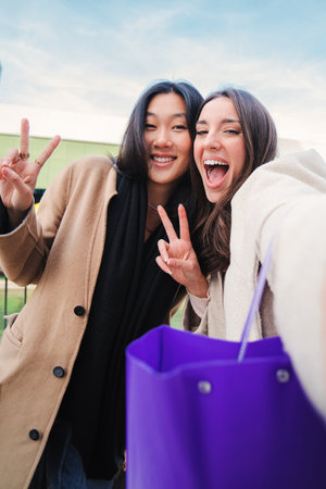 Vertical portrait of two happy young women having fun and laughing taking a selfie photo with smartphone or camera, pretty girls smiling and gesturing the peace sign outsideの写真素材