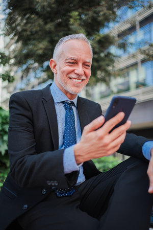 Vertical. Smiling mature businessman with suit using a smartphone sitting outside relaxing and watching apps. Happy professional executive holding a cell phone and reading a