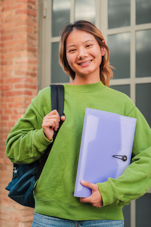 Vertical portrait of a young asian high school student girl smiling and looking at camera. Chinese teenage female staring front at university campus. Thai classmate woman withの写真素材