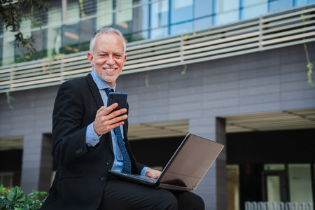 A smiling white-collar worker, wearing a suit and tie, sits on a bench near a buildings facade with a laptop and cellphone. Happy business man using a mobile phone and a computerの写真素材