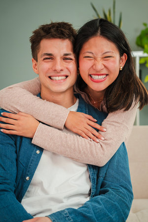 Vertical portrait of a real multiethnic couple smiling, embracing and looking at camera together al...