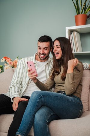 Vertical. Couple having fun using a mobile phone to browse on internet at living room sofa. Two friends betting online with a cellphone app. Boyfriendの写真素材